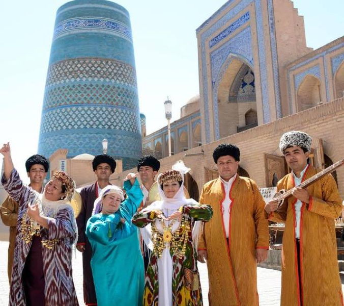 Touristen und Einheimische in traditioneller usbekischer Tracht vor dem blauen Ichan-Kala Minarett in Chiwa.