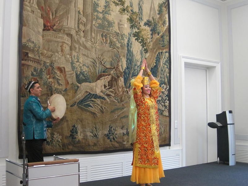 Nawruz 2012 Tänzerin in traditionellem gelb-orangenem Kostüm mit erhobenen Händen vor Wandteppich, Begleiter spielt Rahmentrommel in Raum.