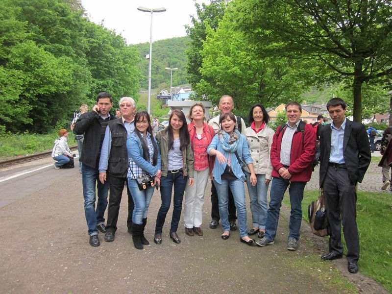 Wanderung mit den usbekischen Studenten im Mai 2012 Gruppe von elf lächelnden Erwachsenen am Bahnhofsvorplatz vor grünen Bäumen und Bahngleisen, Reisende mit Jacken und Rucksäcken