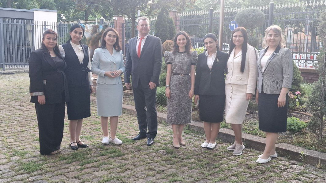 Acht Personen in formeller Kleidung stehen nebeneinander in einem Garten vor einem Zaun, Gruppenfoto bei einer offiziellen Veranstaltung